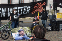 Art Bühne mit Banner "wir machen weiter>" Mikrofon, Foto von Karsten, cola Flasche, kleines Banner "Lieber in Bäumen wohnen statt Wälder roden", Bauwagen aus Pppkarton, Karstens fahrrad. Eine rednerin am Mikrofon