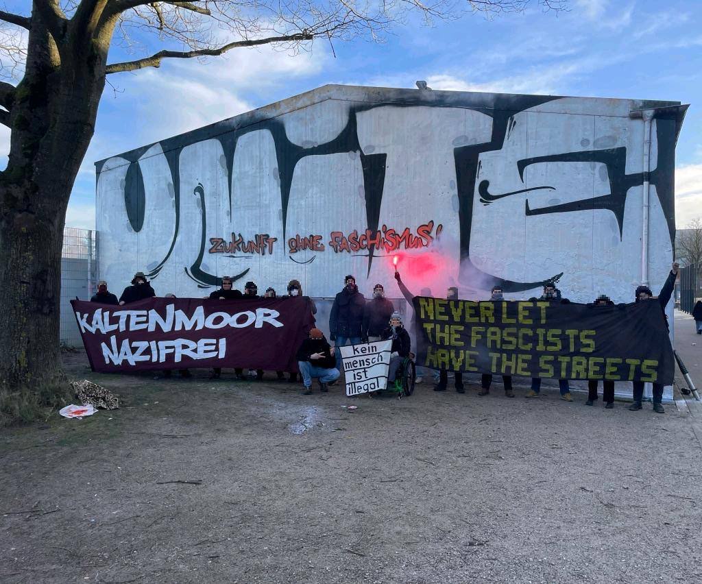 Menschen vor einem Gebaude worauf groß "UNITE" gespayt ist und kleine "Zukunft ohne Faschismus". Davor Menschen mit Banner "Kaltenmoor Nazifrei", "Never let the faschists have the streets, kein Mensch ist illegal"